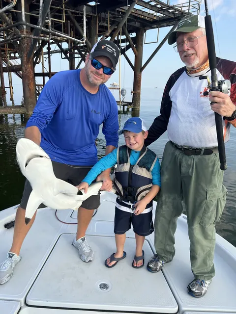 A young angler in a life jacket between two adults, one holding a small shark, on the boat near Venice, Louisiana