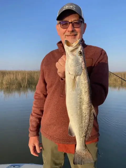Angler in a rust sweater holding up a speckled trout in front of the marsh near Venice, Louisiana